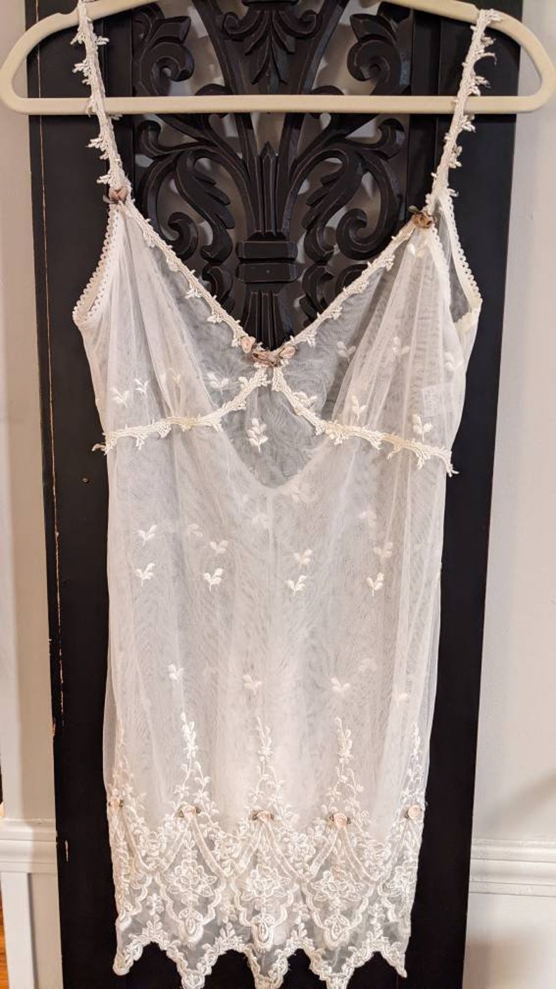 Puede incluir: Un vestido blanco de encaje transparente con delicado bordado floral y ribete de encaje. El vestido tiene un escote redondo y tirantes finos.