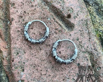 Op de afbeelding: Twee zilveren oorringen met een prikkeldraadontwerp. De oorringen liggen op een bruin, getextureerd oppervlak.