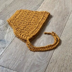 May include: A crocheted baby bonnet in a mustard yellow color. The bonnet has a pointed top and ties that can be tied under the chin.