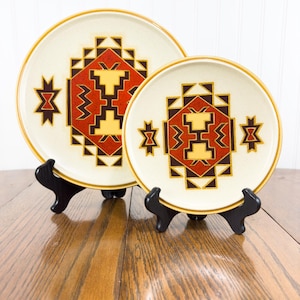 May include: Two ceramic plates with a Southwestern design featuring geometric patterns in shades of red, yellow, and black on a cream background. The plates are displayed on black stands.