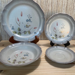 May include: Set of four ceramic plates with a floral design in shades of blue, green, and pink. The plates have a scalloped edge and a light gray background.