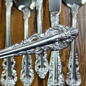 May include: Close-up of ornate silver silverware, including forks and a knife, with detailed floral and scrollwork patterns. The silverware is arranged on a wooden surface, showcasing the intricate designs and polished finish.