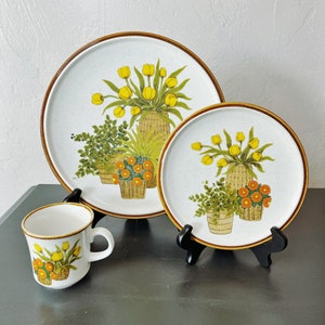 May include: A set of three white ceramic plates with a brown rim and a floral design featuring yellow tulips, green plants, and orange flowers in brown baskets.