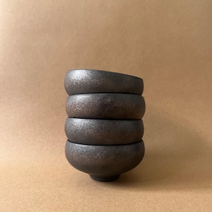 May include: A stack of four small, dark brown ceramic bowls. The bowls have a rounded shape with a slightly textured surface. The bowls are stacked on top of each other, with the bottom bowl resting on a neutral-colored surface.