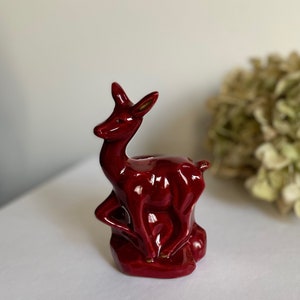 Peut inclure: Une figurine de cerf en céramique de couleur bordeaux avec une finition brillante. Le cerf est debout sur une base et a la tête tournée sur le côté.