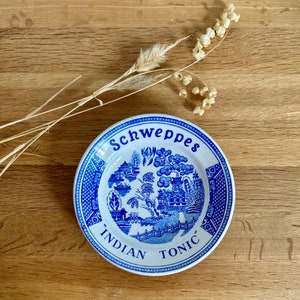 Peut inclure: Une soucoupe en céramique bleue et blanche avec un motif floral bleu et blanc et le texte "Schweppes Indian Tonic" dans un cercle blanc.