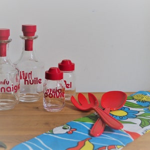 Peut inclure: Quatre bouteilles en verre avec des bouchons et des étiquettes rouges. Les bouteilles sont étiquetées "vinaigre", "huile", "poivre" et "sel". Les bouteilles sont posées sur une table en bois avec un tissu à motifs bleus et jaunes devant elles. Il y a un service à salade en plastique rouge sur le tissu.