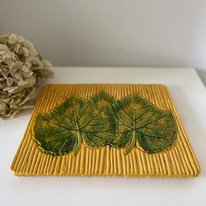 Peut inclure: Un carreau de céramique carré avec un motif en relief de trois feuilles vertes sur fond jaune. Les feuilles ont un motif de nervures détaillé et sont disposées sur un fond texturé qui ressemble à du bambou.