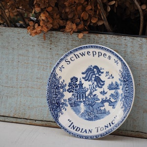 Peut inclure: Une assiette décorative blanche et bleue avec le texte "Schweppes" et "Indian Tonic". L'assiette présente une illustration bleue détaillée d'une pagode, d'arbres et d'un pont. L'assiette est ronde et a une bordure bleue à motifs.