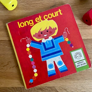 Peut inclure: Un livre rouge avec le titre "long et court" et une illustration de dessin animé d'un enfant portant une chemise et un pantalon bleus. Le livre fait partie d'une collection appelée "pré-école".