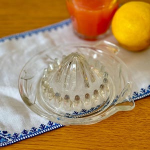 Peut inclure: Un presse-agrumes en verre transparent avec un centre strié et une base large. Le presse-agrumes est posé sur un tissu blanc avec des broderies bleues.
