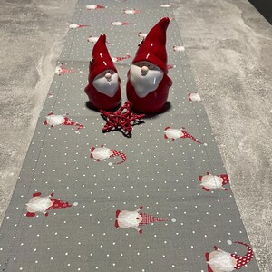 Könnte beinhalten: Grauer Tischläufer mit einem sich wiederholenden Muster aus weißen Punkten und Zwergenfiguren in Rot und Weiß. Zwei rote Zwergfiguren und eine rote sternförmige Dekoration liegen darauf. Festliches Urlaubsthema.