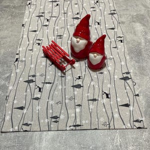 Könnte beinhalten: Ein grauer und weißer Tischläufer mit einem winterlichen Design mit schwarzen Bäumen, Schneeflocken und wellenförmigen Linien. Zwei rote Keramik-Zwerge sind auf dem Tischläufer platziert, und ein roter Holzschlitten befindet sich im Vordergrund.