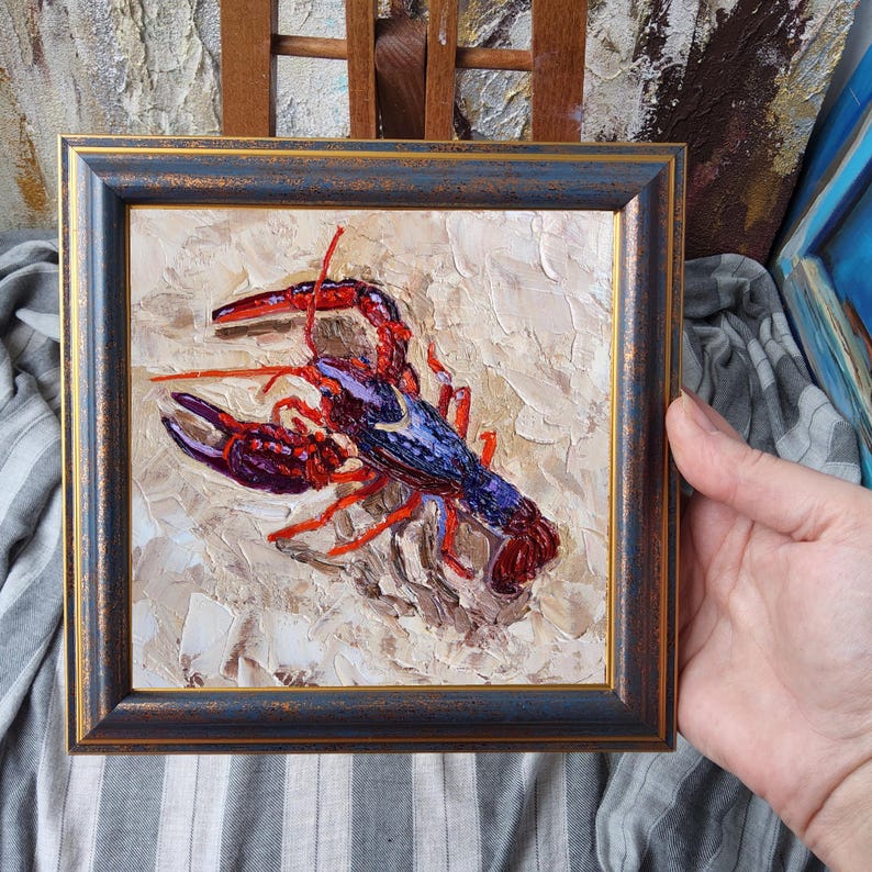 May include: Square framed oil painting of a lobster. The lobster is painted with red, purple, and blue hues, set against a textured beige background. The frame is dark blue with gold trim.