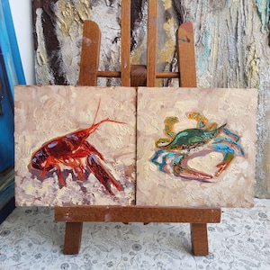 May include: Two small square oil paintings on a wooden easel. One painting depicts a red crawfish, the other a blue and green crab. Both paintings have a textured, impasto style with a neutral background.