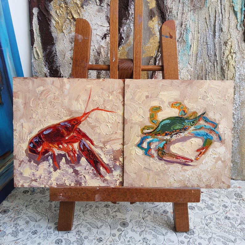 May include: Two small square oil paintings on a wooden easel. One painting depicts a red crawfish, the other a blue and green crab. Both paintings have a textured, impasto style with a neutral background.