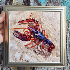 May include: Oil painting of a lobster in a gold frame. The lobster is painted with shades of red, purple, and blue, set against a textured beige background. The artwork is a square format.