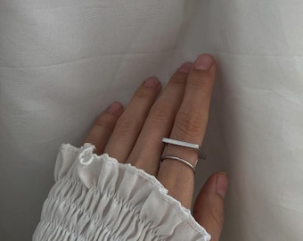 May include: A silver ring with a square design. The ring is worn on the middle finger of a hand.