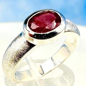 May include: A silver ring with a red oval gemstone set in a bezel setting. The ring band has a textured finish.