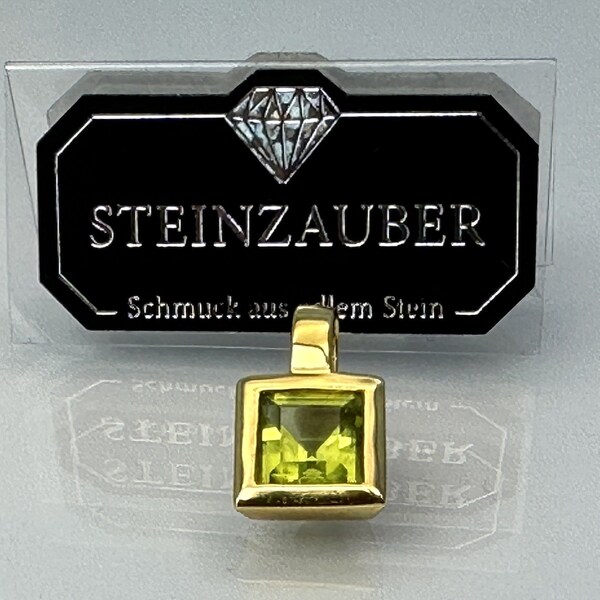 May include: A gold pendant with a square green gemstone. The pendant is on a white surface with a black and white sign that reads "STEINZAUBER Schmuck aus dem Stein".