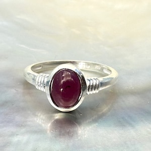 Könnte beinhalten: Ein silberner Ring mit einem tief roten ovalen Edelstein, der in einer Zargenfassung sitzt. Der Ringarm ist mit Silberdraht umwickelt.