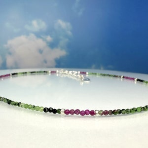 Könnte beinhalten: Eine zarte Halskette aus kleinen grünen, rosa und silbernen Perlen. Die Halskette liegt auf einer weißen Oberfläche.