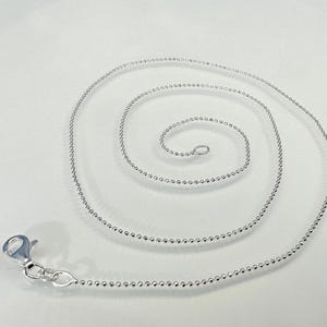 Op de afbeelding: Een zilverkleurige kogelketting met een karabijnsluiting, opgerold in een spiraal op een witte ondergrond. De ketting is gemaakt van kleine, ronde kralen, wat een delicate en elegante uitstraling creëert. De sluiting is open.