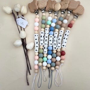 Op de afbeelding: Zeven siliconen-bijtkettingen met houten clip-speeltjes. De kettingen zijn gemaakt van verschillende gekleurde kralen en hebben gepersonaliseerde letterkralen die namen spellen zoals "Pinky", "Kiwi", "Twinkle", "Sky", "Moon", "Teddy" en "Berry".