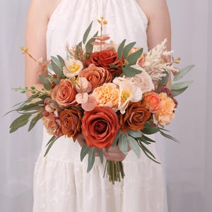 May include: A bouquet of orange, peach, and cream coloured artificial flowers with green leaves. The bouquet is held by a person wearing a white dress.