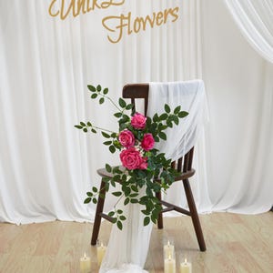 May include: A wooden chair adorned with pink roses and green foliage, draped with white fabric. The scene is set against a backdrop of white curtains and includes candles. The text "Unik Love Flowers" is visible.
