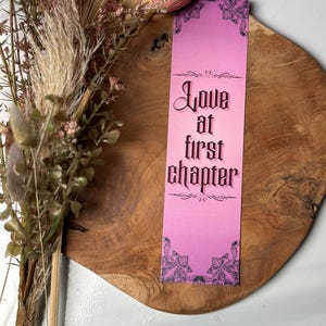 May include: Pink bookmark with black text that reads "Love at first chapter" with a decorative black border.