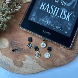 May include: A Kindle e-reader showing the book "Kiss of the Basilisk" by Lindsay Straube, with four decorative charms featuring celestial and animal designs. The charms are black, gold, and white, on a wooden tray.