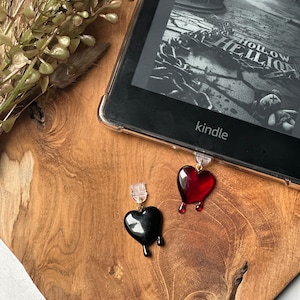 May include: Two heart-shaped book markers, one black and one red, with dripping edges. The markers are attached to a Kindle e-reader with the words "Hollow" and "The" visible on the screen.