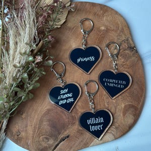 Op de afbeelding: Hartvormige sleutelhangers met zilveren sleutelringen. Elke zwarte sleutelhanger heeft witte tekst: "princess", "such a f*cking good girl", "villain lover" en "completely unhinged". De sleutelhangers worden op een houten oppervlak gepresenteerd.