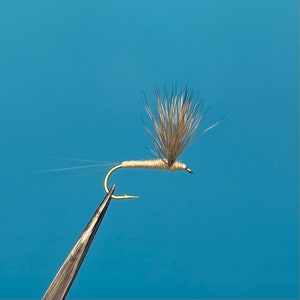 May include: A light brown dry fly fishing lure with a gold hook and a white body. The fly has a long tail and a bushy wing.