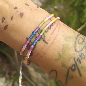 Può includere: Un polso con un braccialetto di corda colorato. Il braccialetto è fatto di più fili di corda di diversi colori, tra cui rosa, blu, giallo e verde. La persona ha diversi tatuaggi sul braccio, tra cui un fiore, un sole e un uccello.