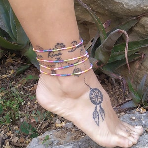 Puede incluir: Primer plano de un tobillo con un tobillo de cuentas de colores y un tatuaje de atrapasueños. El tobillo está hecho de pequeñas cuentas de colores y está atado alrededor del tobillo. El tatuaje de atrapasueños es en blanco y negro y está ubicado en el tobillo.