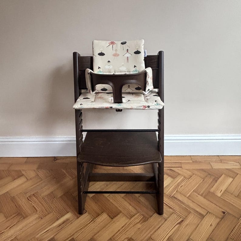 May include: A dark brown wooden high chair featuring a removable dark brown plastic safety rail and a padded seat cushion set. The cushion fabric is cream-colored and decorated with a repeating pattern of small, stylized ballerinas in black, pink, and teal.