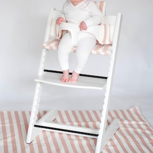 Pu&ograve; includere: Un seggiolone in legno bianco dotato di una barra di sicurezza in plastica bianca e un morbido cuscino per la seduta con un motivo a righe rosa chiaro e bianco. La sedia presenta livelli regolabili per sedile e poggiapiedi, offrendo stabilit&agrave;.