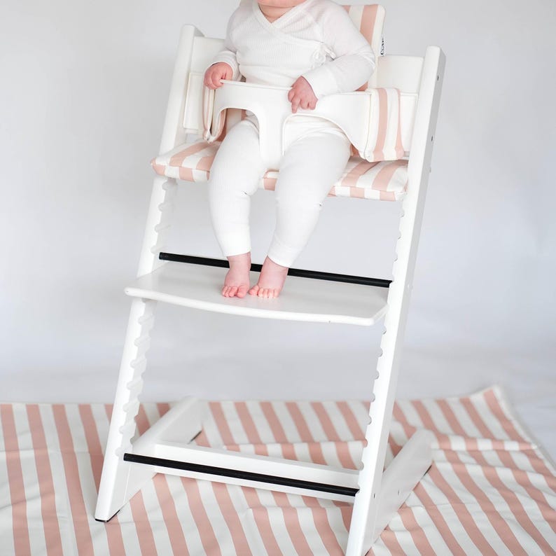 Pu&ograve; includere: Un seggiolone in legno bianco dotato di una barra di sicurezza in plastica bianca e un morbido cuscino per la seduta con un motivo a righe rosa chiaro e bianco. La sedia presenta livelli regolabili per sedile e poggiapiedi, offrendo stabilit&agrave;.