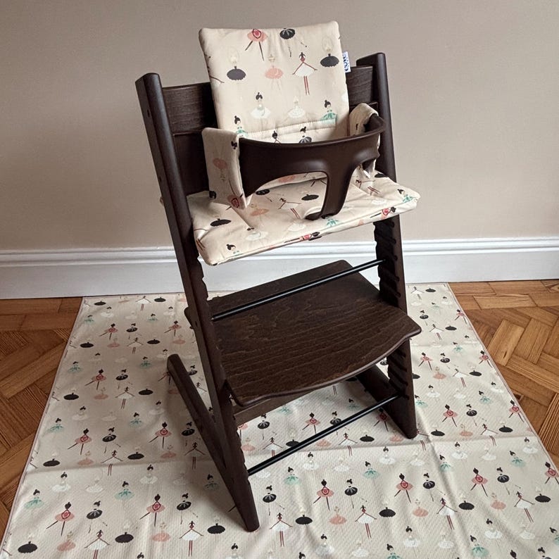 May include: A dark brown wooden high chair featuring a beige seat cushion and backrest pad decorated with a repeating pattern of small ballerinas in black, pink, and teal. The chair includes a dark brown plastic safety rail and footrest.