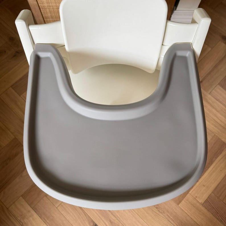 May include: Overhead view of a white high chair with a gray tray. The chair has a curved back and armrests. The tray is a light gray color and is attached to the front of the chair.
