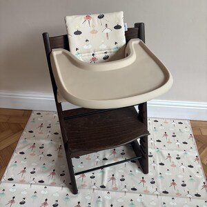 May include: A dark brown wooden high chair featuring a beige plastic tray and a cushioned backrest with a repeating pattern of stylized ballerinas in black, white, pink, and teal. A matching fabric mat with the same ballerina print is underneath.