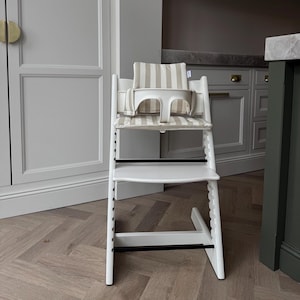 May include: A white wooden high chair with a striped beige and white cushion and backrest. The chair has a footrest and a safety bar. The chair is designed for a baby or toddler.