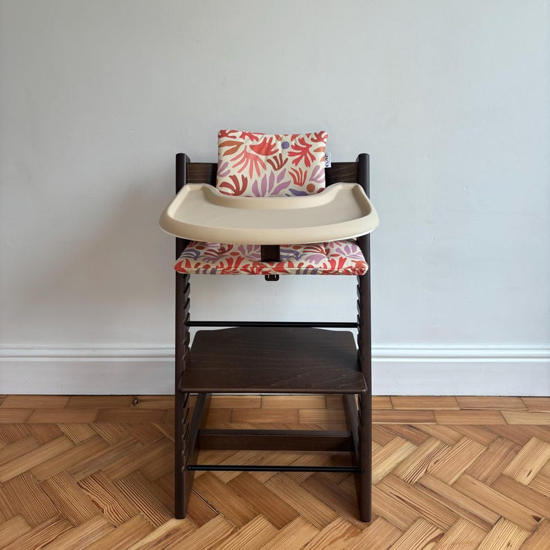 May include: A dark brown wooden high chair featuring a beige plastic tray and a padded seat cushion with a colourful abstract botanical print in shades of red, pink, and purple. The chair includes a matching backrest cushion and a footrest.