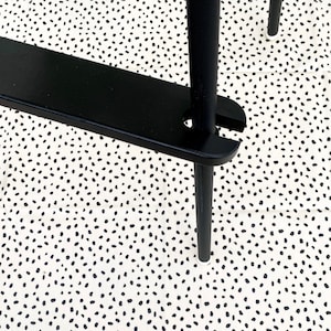 A black wooden footrest that is fitted on a toddlers  high chair legs. Accompanied with black leg wraps and on top of a black spotted pink splash mat.