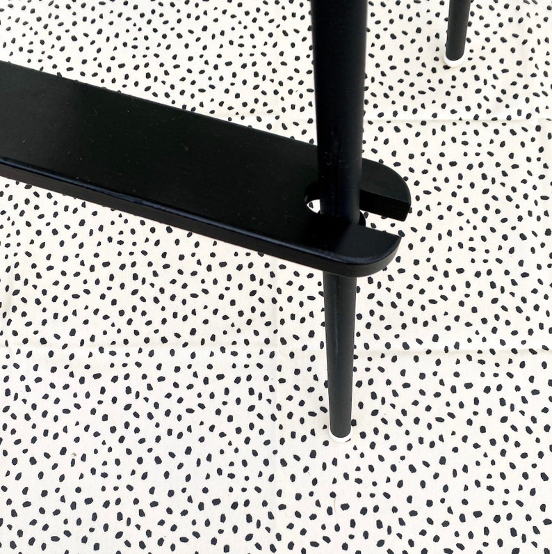 A black wooden footrest that is fitted on a toddlers  high chair legs. Accompanied with black leg wraps and on top of a black spotted pink splash mat.