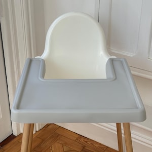 May include: A modern high chair with a white seat and a gray tray. The chair has light-colored wooden legs. The tray is rectangular and has raised edges. The chair is designed for a baby or toddler.