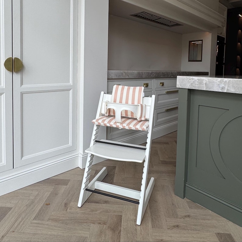 Pu&ograve; includere: Un seggiolone in legno bianco con cuscino per seduta e schienale a righe rosa chiaro e bianco. La sedia include una barra di sicurezza in plastica bianca, un poggiapiedi e una seduta regolabile in altezza tramite le fessure laterali.