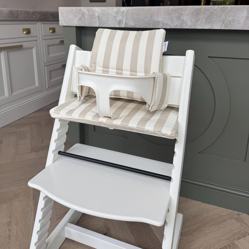 May include: A white wooden high chair with a beige and white striped cushion and backrest. The high chair has a white plastic safety bar and a black footrest. The chair is designed for infants and toddlers.